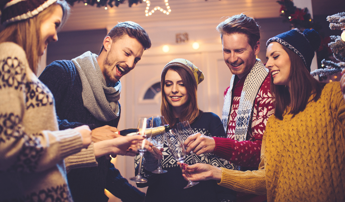 Young group of friends having fun outdoors in front of house. Pouring champagne in glasses. Wearing knitted sweaters, hats and scarfs. House, yard and tree are decorated with festive string lights. Evening or night with beautiful yellow lights lightning the scenes.